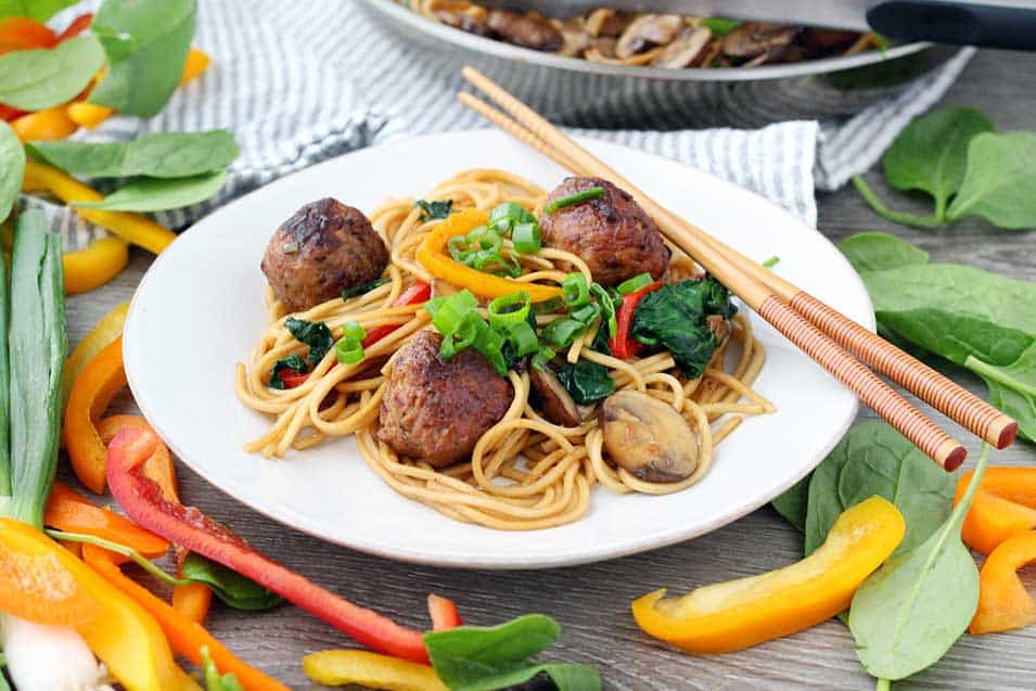 A white plate with meatball lo mein, veggies around it, and chopsticks.