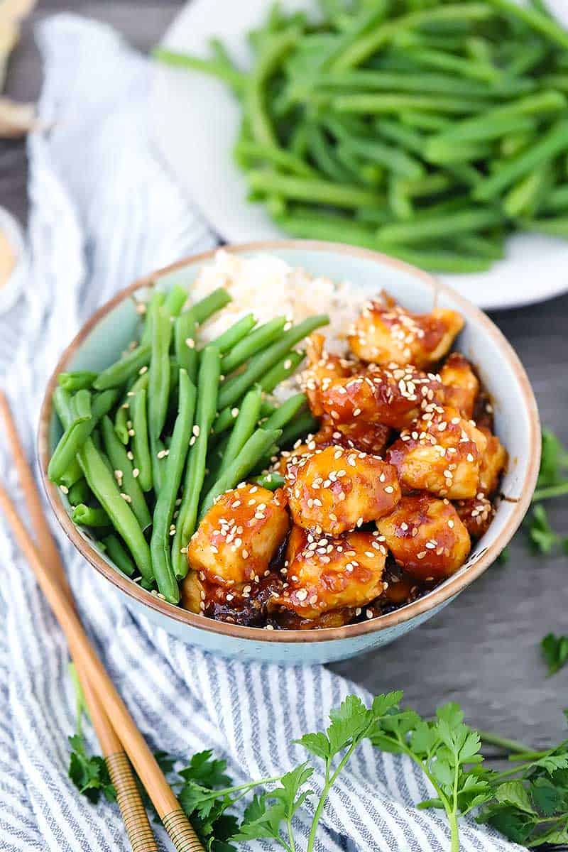 A bowl with sesame chicken, green beans, and rice with chopsticks on the side.