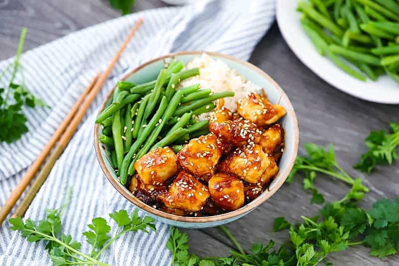 Horizontal photo of a bowl of sesame chicken with green beans and rice.