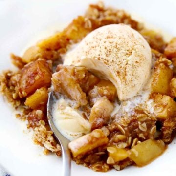 Apple crisp with vanilla ice cream melting on top and a spoon taking a bite out of it.