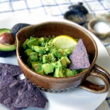 Avocado salsa in a brown ceramic bowl with handle, surrounded by tortilla chips and avocados.