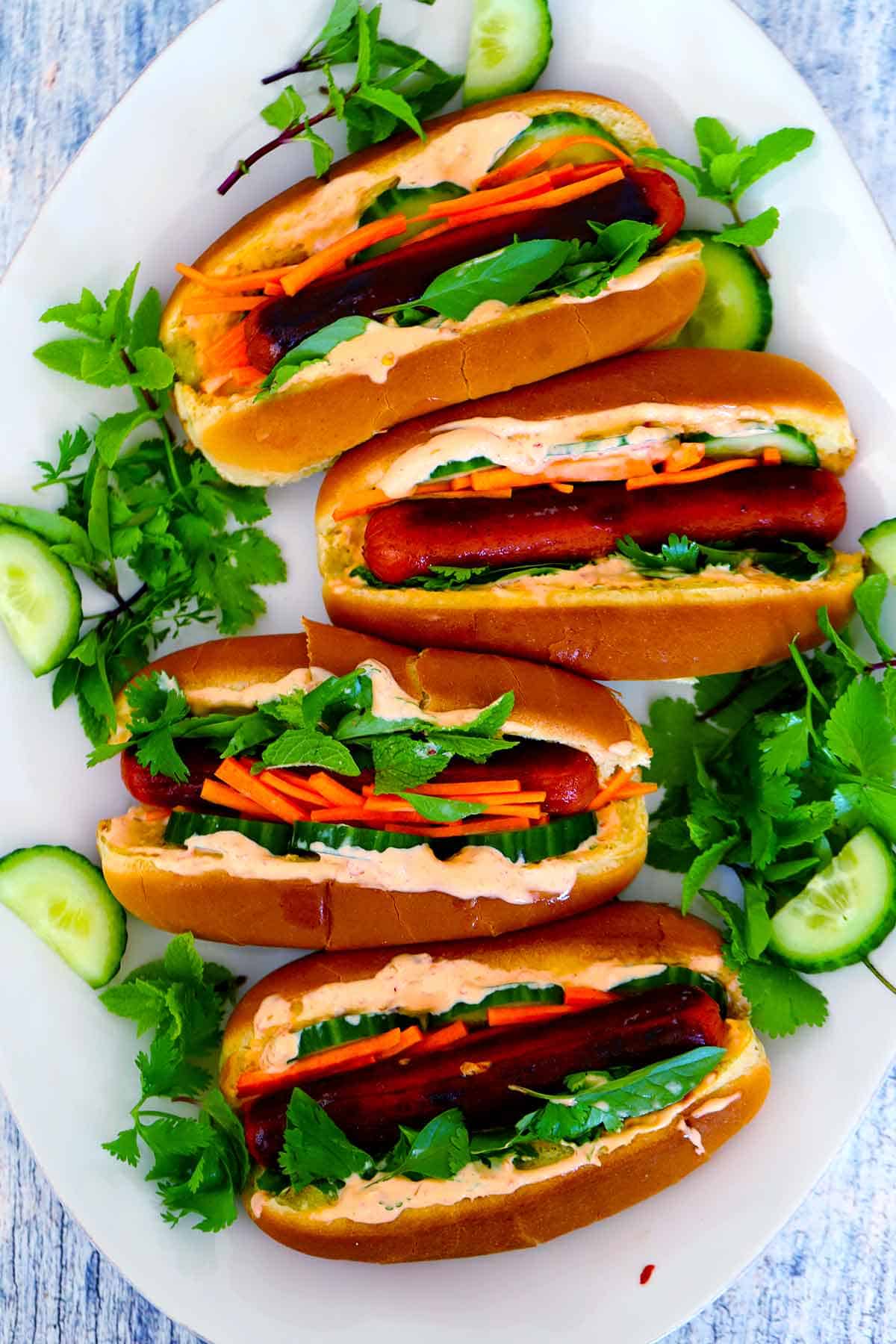 Four Bánh Mì hot dogs on a large white platter, overhead view, surrounded by fresh herbs and cucumber slices.