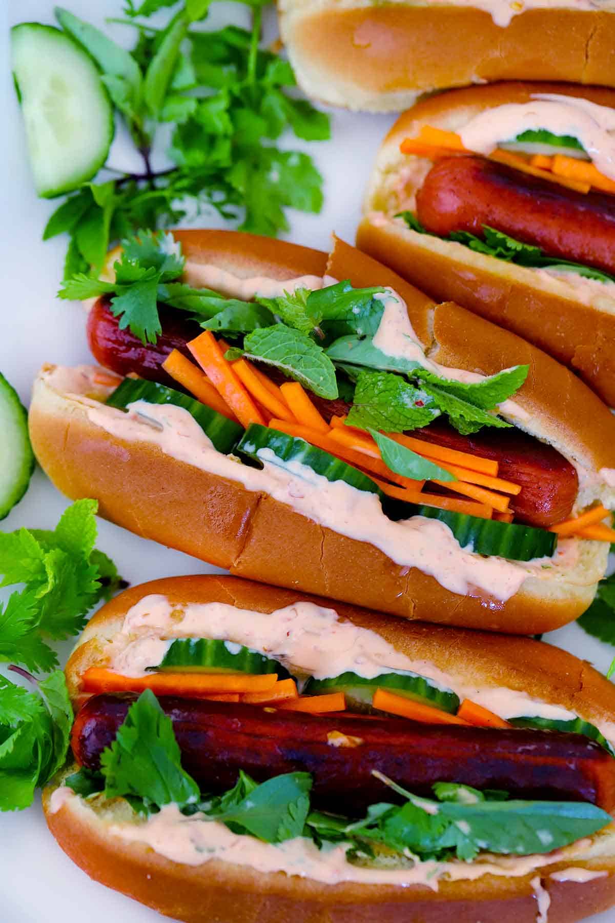Close up overhead view of Bánh Mì hot dogs, with spicy mayo, pickled carrots, cucumbers, and herbs.