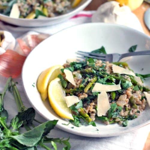A white plate holding barley risotto with asparagus, basil, and lemon, as well as thin parmesan slices, is in the foreground. Another bowl of the dish, as well as ingredients, are scattered in the background and around the foreground bowl.