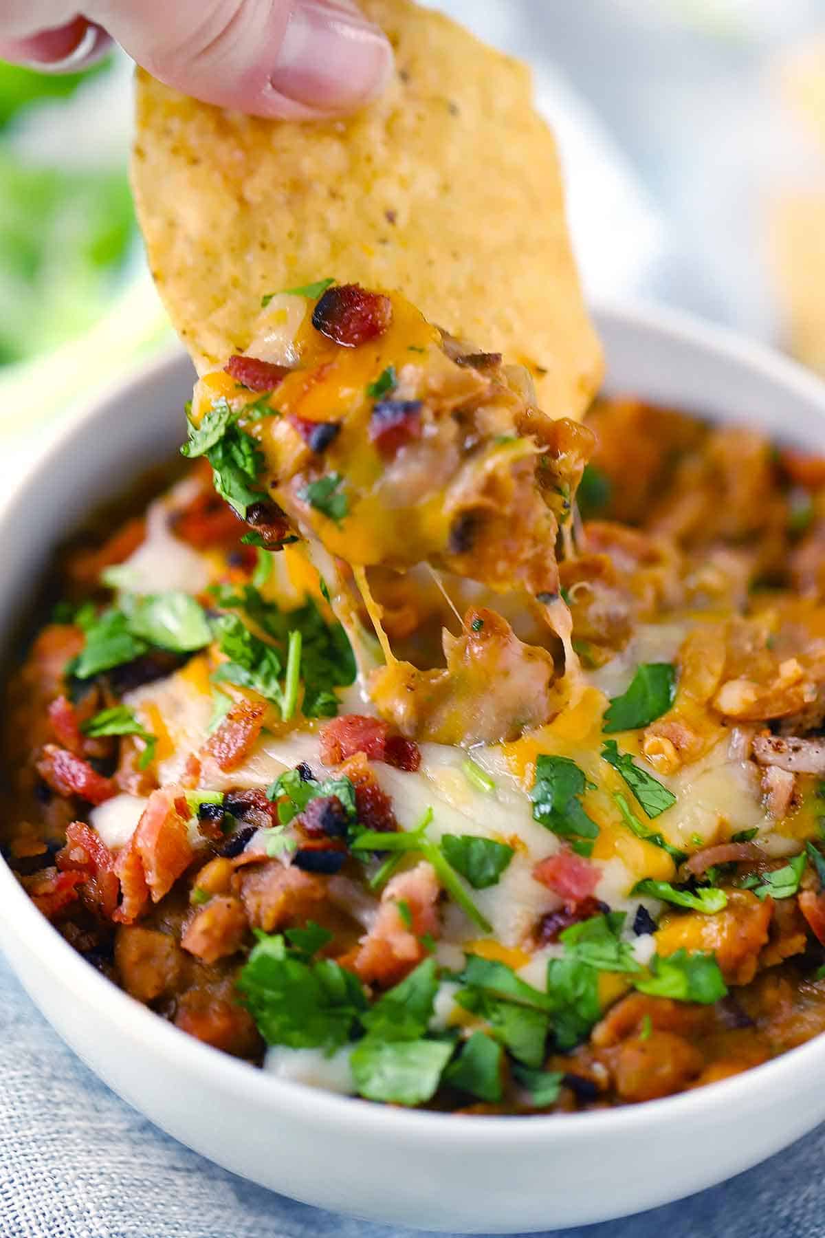 A tortilla chip being dipped in a bowl of refried beans with melted cheese on top.