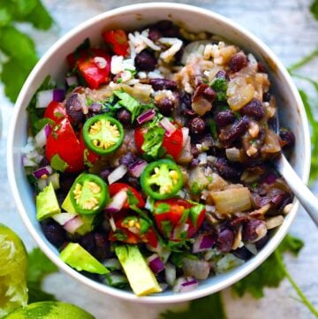Over head photo of a bowl of black beans and rice topped with jalapeños, tomatoes, and onion, surrounded by cilantro and lime.