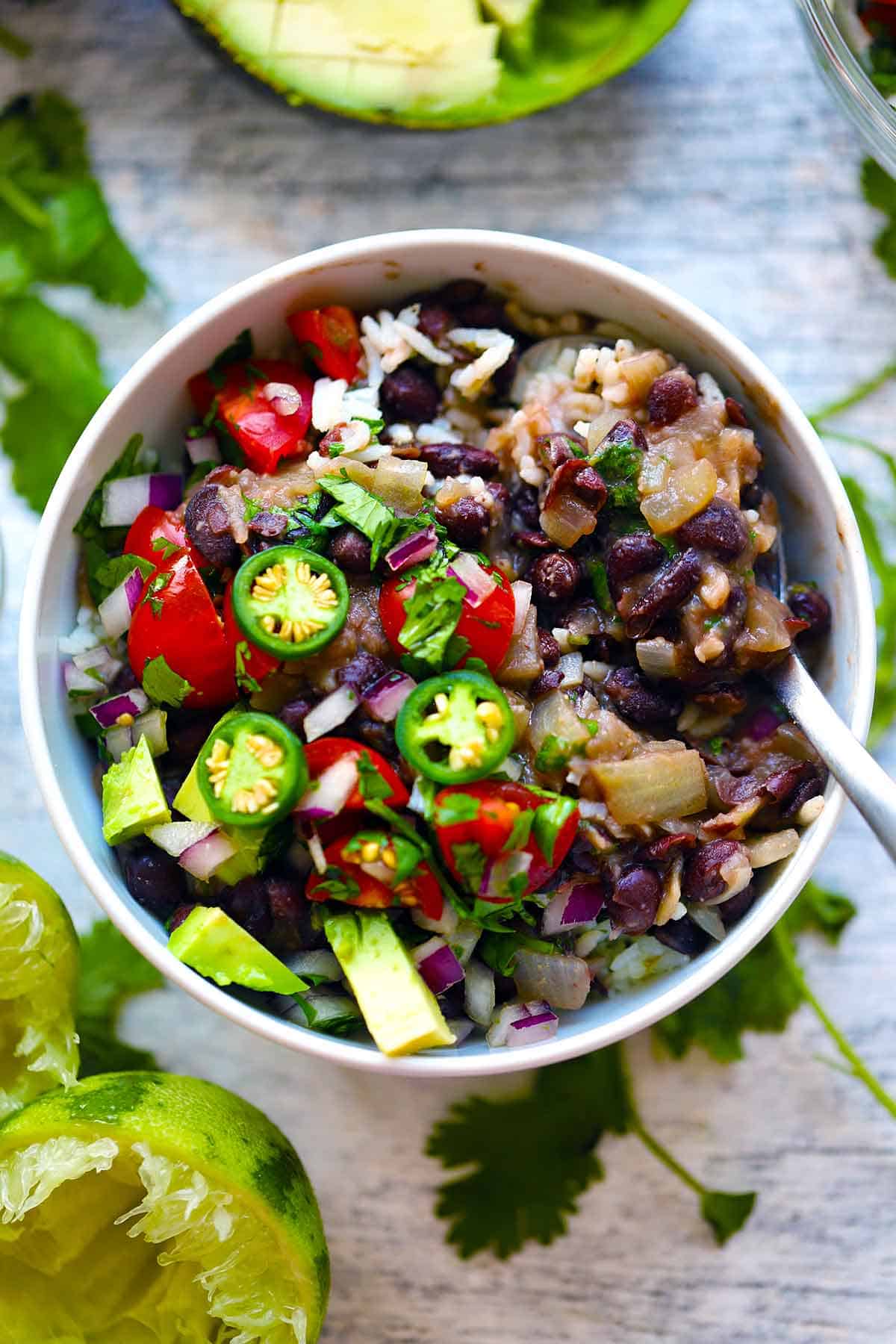 Over head photo of a bowl of black beans and rice topped with jalape&ntilde;os, tomatoes, and onion, surrounded by cilantro and lime.