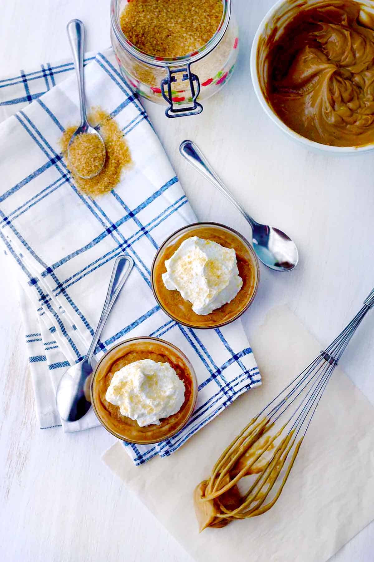Overheat photo of two bowls of homemade butterscotch pudding with whipped cream, spoons and whisk around them.