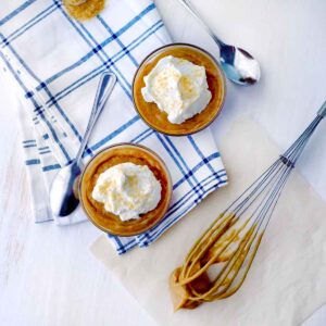 Square photo of two bowls of butterscotch pudding and a whisk on the side.