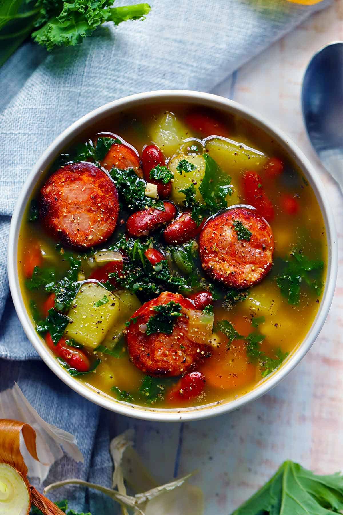 Overhead photo of caldo verde soup showing sausages, beans, kale, and potatoes in a white bowl.