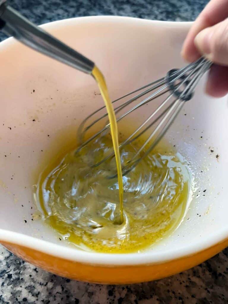 Mixing a vinaigrette in a vintage pyrex bowl using a whisk and drizzling olive oil in.