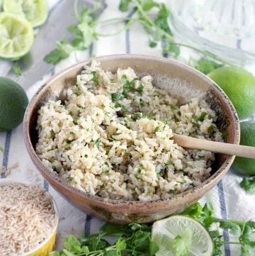 Brown rice in brown ceramic bowl, with wooden spoon and limes and parsley around bowl.