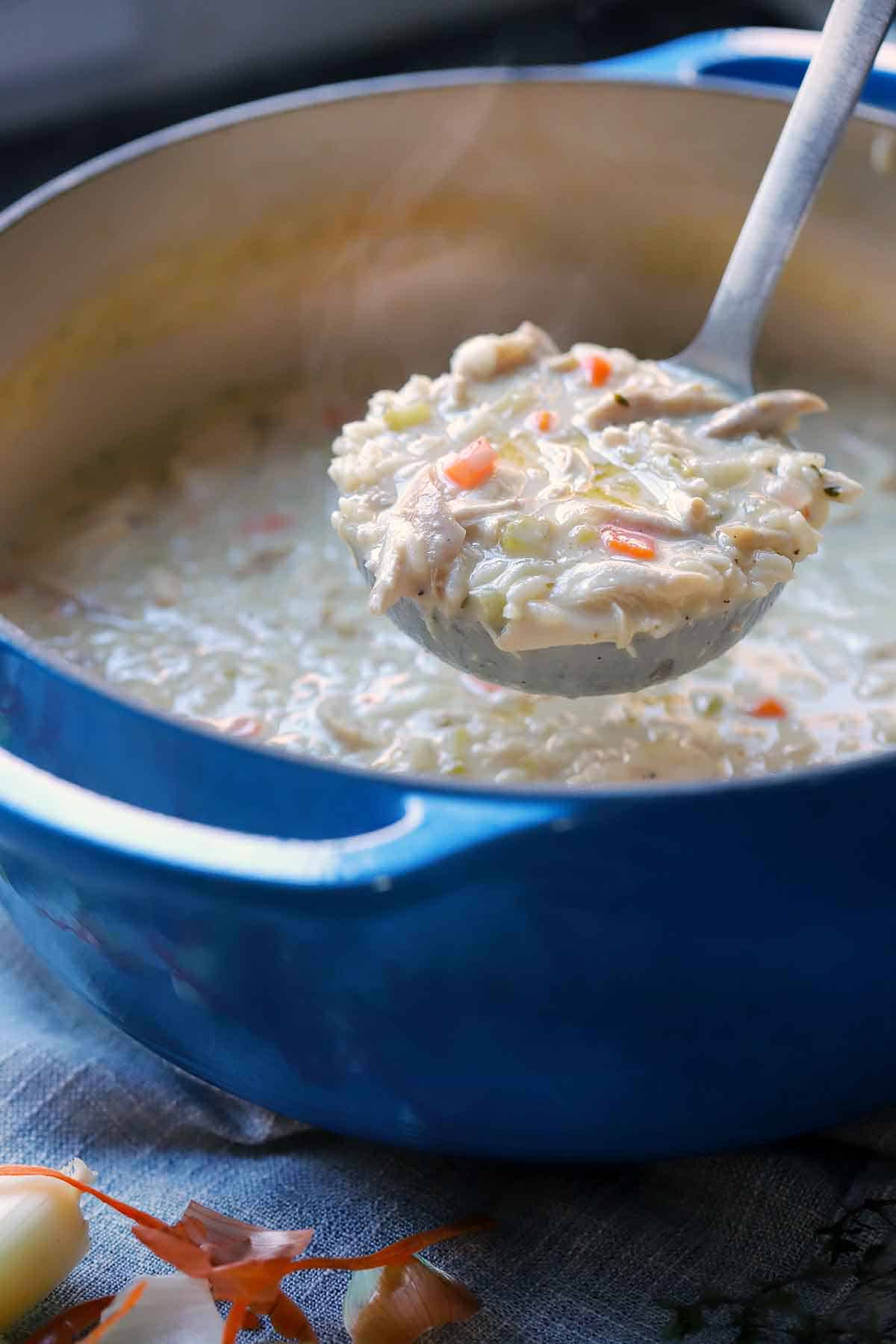 A ladle lifting a scoop of chicken rice soup out of a blue dutch oven, with steam coming from the top.