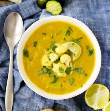 A bowl of curried cauliflower soup on a blue towel with a spoon, garnished with cauliflower florets and grated zucchini.