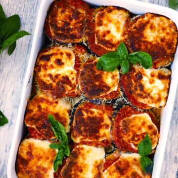 Overhead photo of a white baking dish with eggplant parmesan in it and fresh basil as a garnish with browned crispy cheese on top.