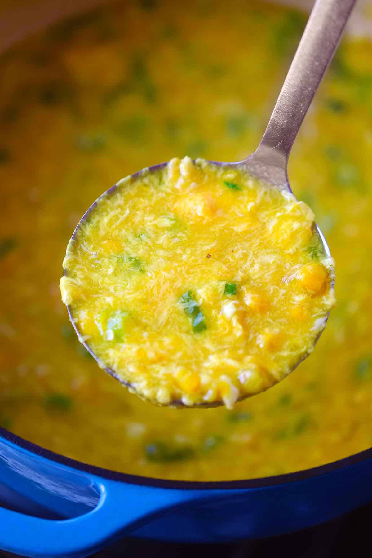 A ladle full of egg drop soup with corn over a blue Dutch oven.