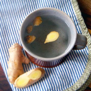 Bird's eye view of ceramic mug with tea inside, with fresh ginger root pieces floating in the tea and a few pieces scattered on the side of the mug.