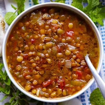 A square photo of Greek lentil soup with a spoon in it, surrounded by herbs.