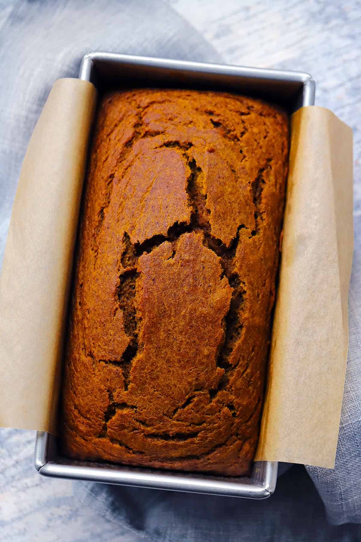 A loaf of pumpkin bread made with whole wheat flour, maple syrup, and olive oil in a loaf pan after baking, with parchment paper on the sides for lifting out,