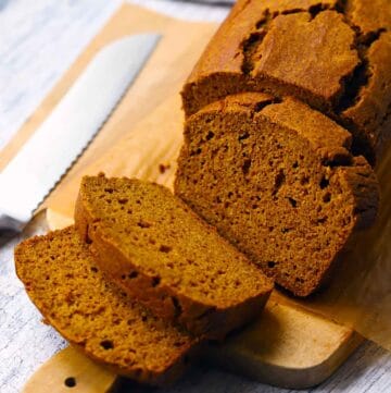 Square photo of a loaf of whole wheat pumpkin bread, sliced, on a wooden cutting board with a bread knife next to it.