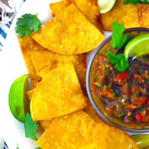Cropped square photo of a plate of homemade tortilla chips, a bowl of salsa, and lime and cilantro.