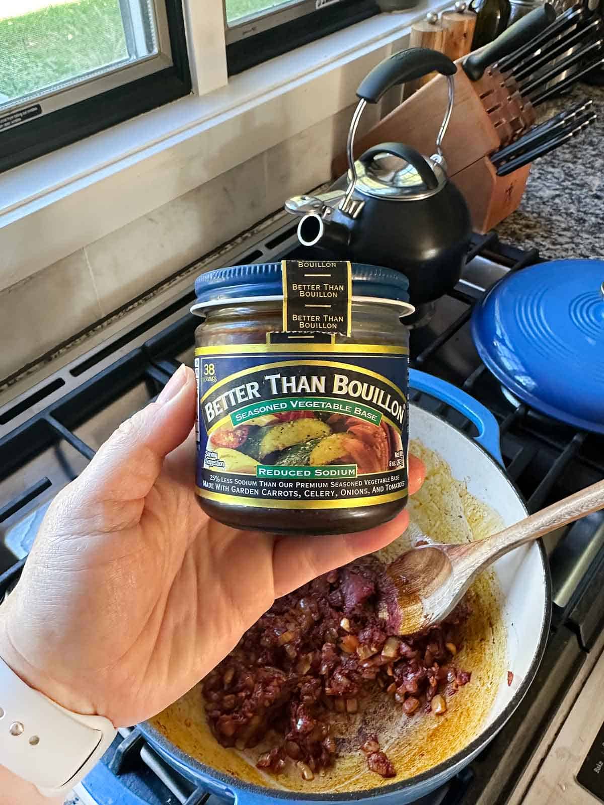 A hand holding a jar of better than bouillon seasoned vegetable low sodium base over a soup pot on the stove.