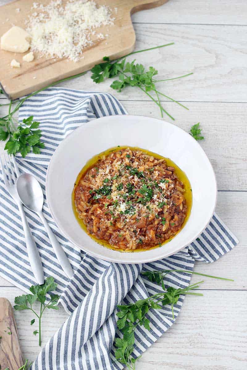 Orzo and ground beef (manestra) in white bowl, on blue and white striped towel, with herb garnish.