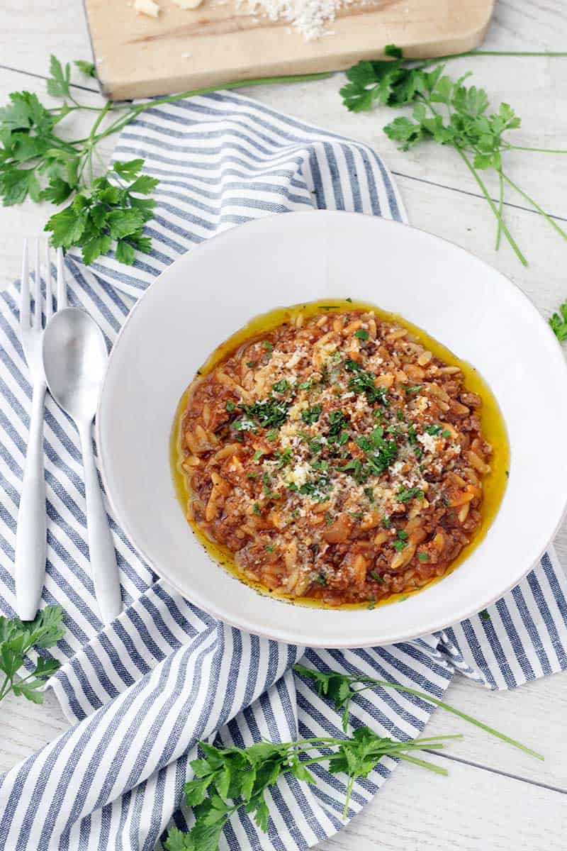 Orzo and ground beef (manestra) in white bowl, on blue and white striped towel, with herb garnish.