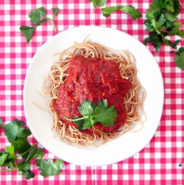 Top down view of pasta with marinara sauce white plate on red and white checkered tablecloth, sprinkled with torn basil.