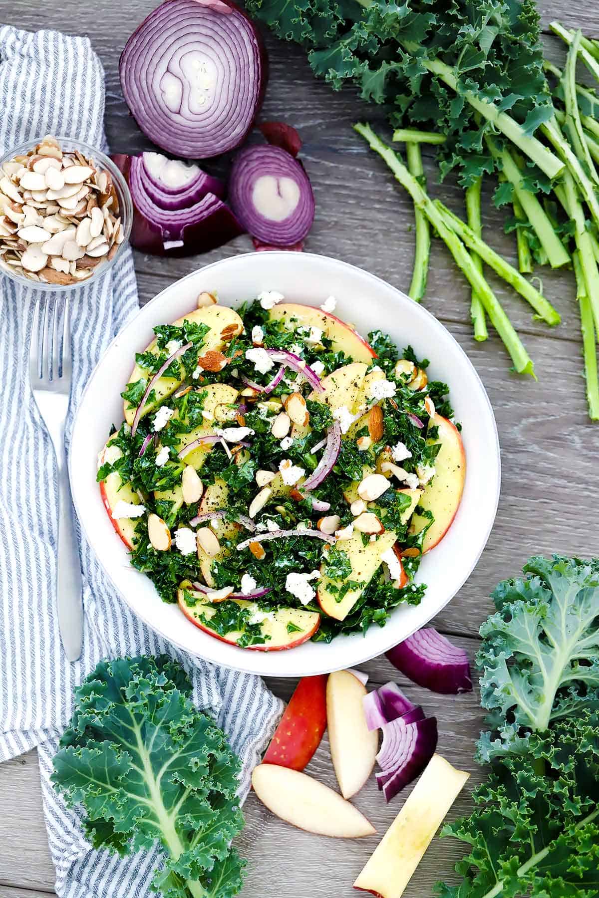 A white plate with kale salad and apples, onions, almonds, a striped towel, and kale scraps scattered around it.