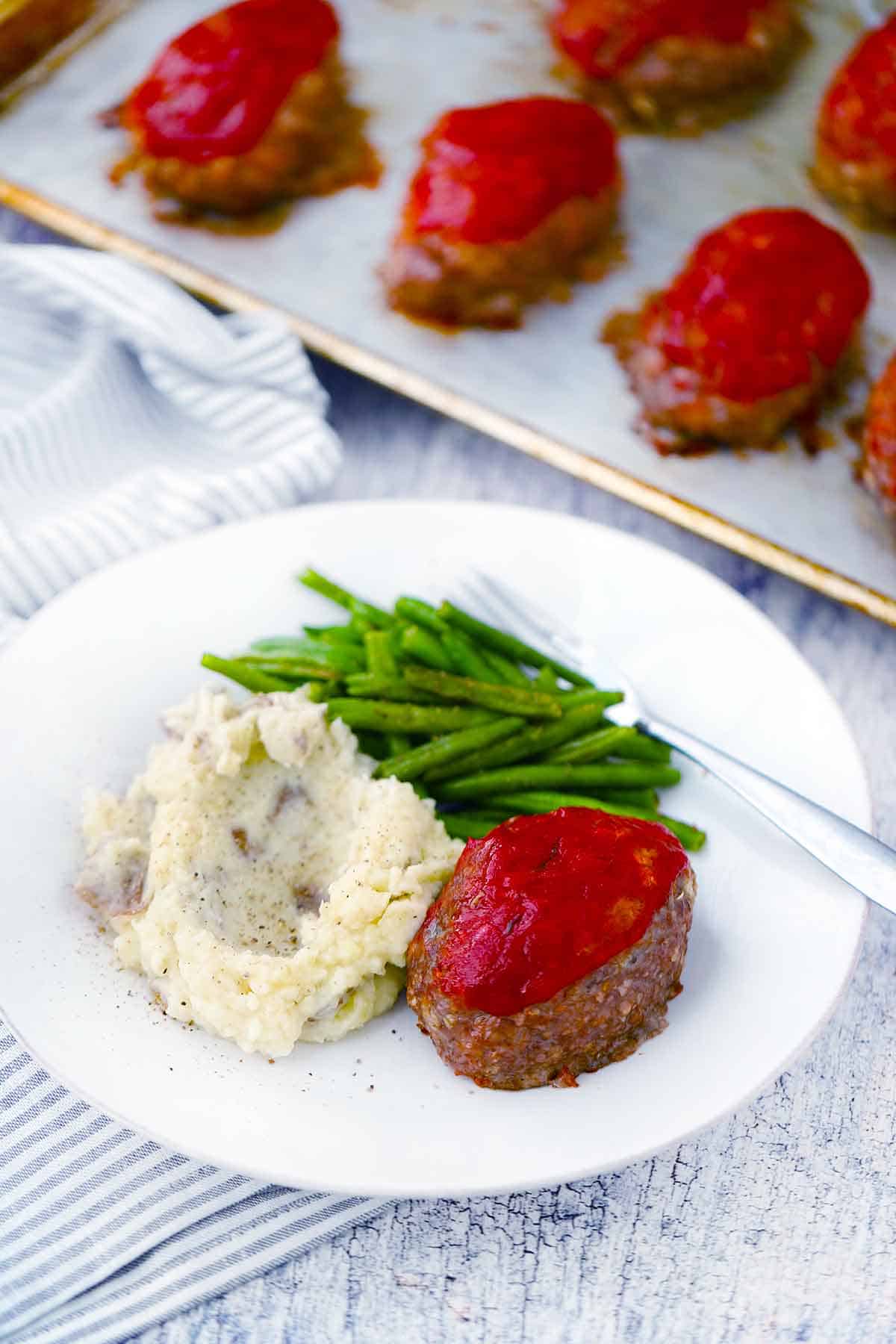 A white plate with a mini meatloaf, green beans, and potatoes, with a baking sheet with more meatloaves.