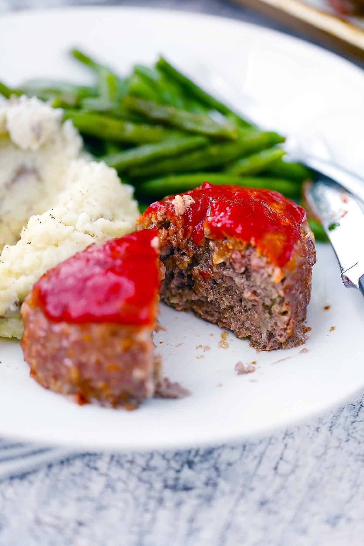 A white plate with a mini meatloaf sliced in half with green beans and mashed potatoes.