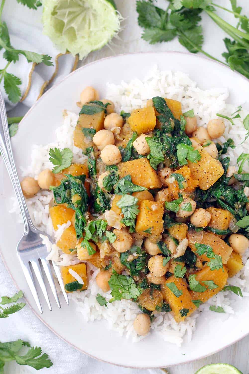 Close up, bird's eye view of Moroccan butternut squash and chickpea stew on a bed of white rise, on a white plate, with a fork perched on the side and herbs scattered around.