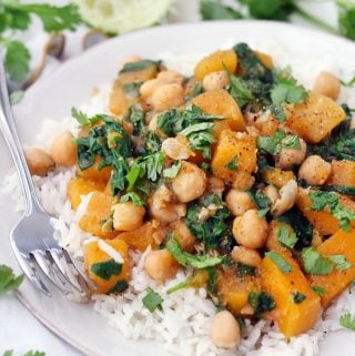 Side view of moroccan butternut squash and chickpea stew on a bed of white rise, on a white plate, with a fork perched on the side and herbs scattered around.