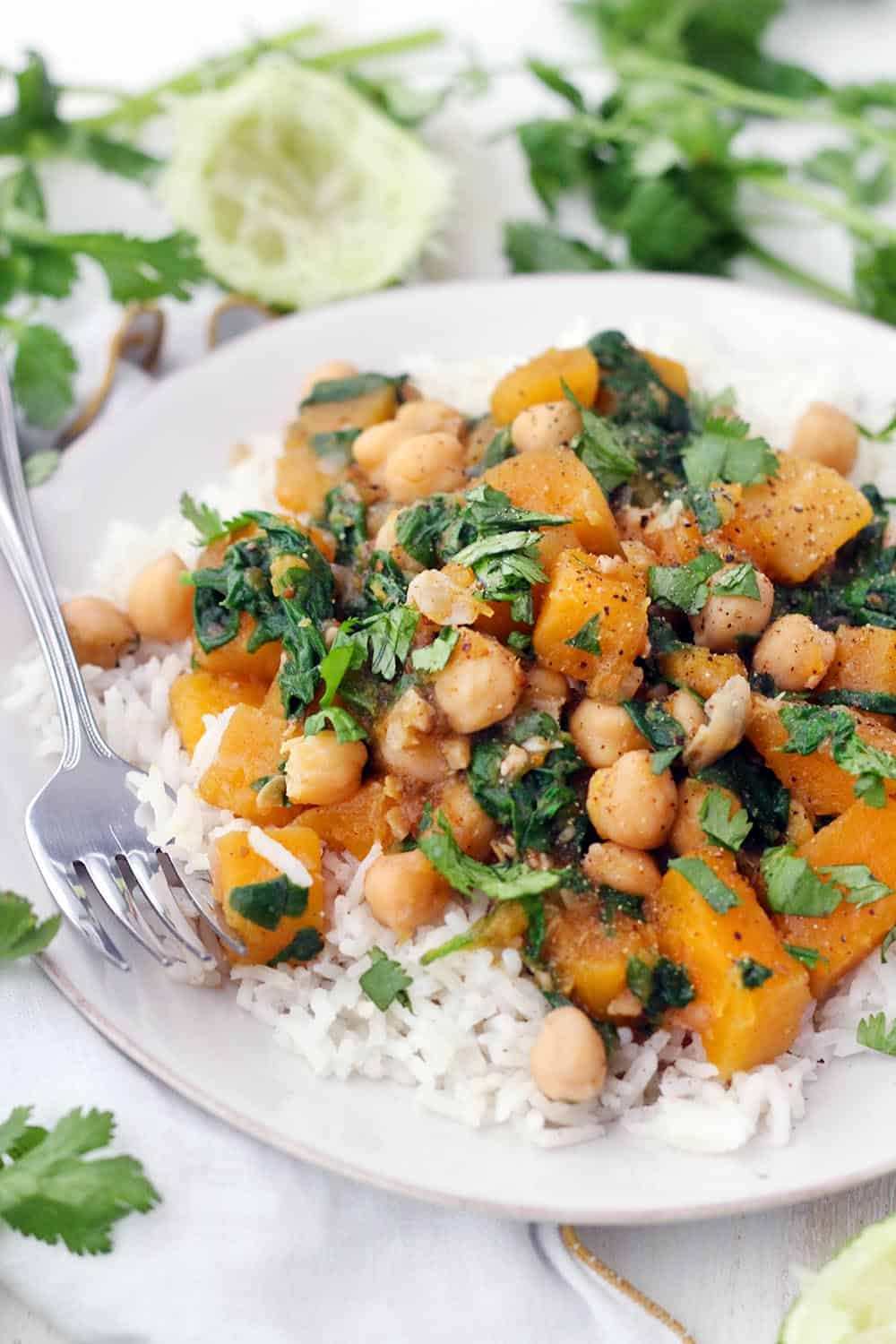 Side view of moroccan butternut squash and chickpea stew on a bed of white rise, on a white plate, with a fork perched on the side and herbs scattered around.