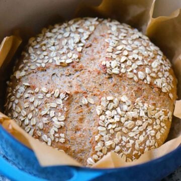 Close up square photo of a seeded multigrain no knead bread loaf after baking in a blue Dutch oven lined with parchment paper.