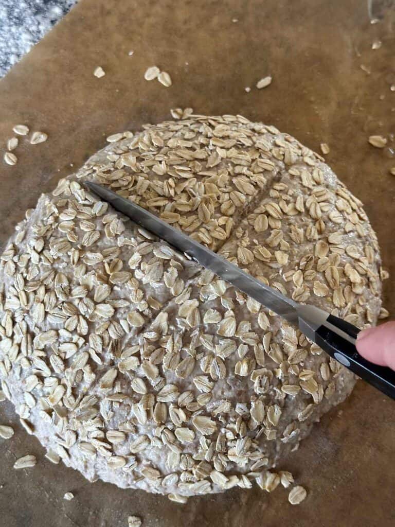 Scoring the top of a whole grain no knead bread loaf with a sharp knife into an X shape.