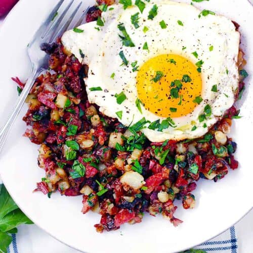 Square photo of corned beef hash on a plate with a fork and a fried egg on top.