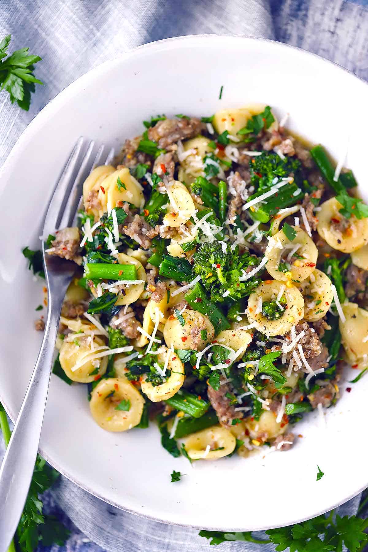 Overhead photo of a white bowl filled with orecchiette, broccolini, and sausage garnished with parmesan with a fork.