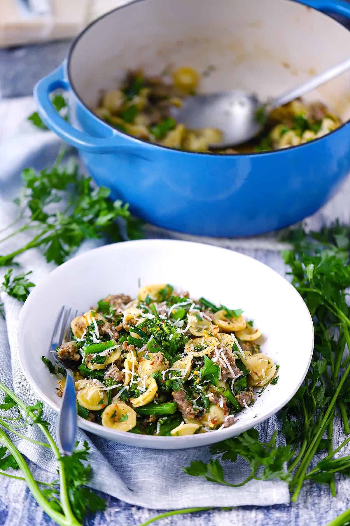 A white bowl filled with pasta with sausage and broccoli and a blue Dutch oven in the background.