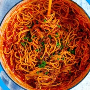 Square photo of an overhead view of a pot with spaghetti and meat sauce garnished with parsley in a blue Dutch oven.