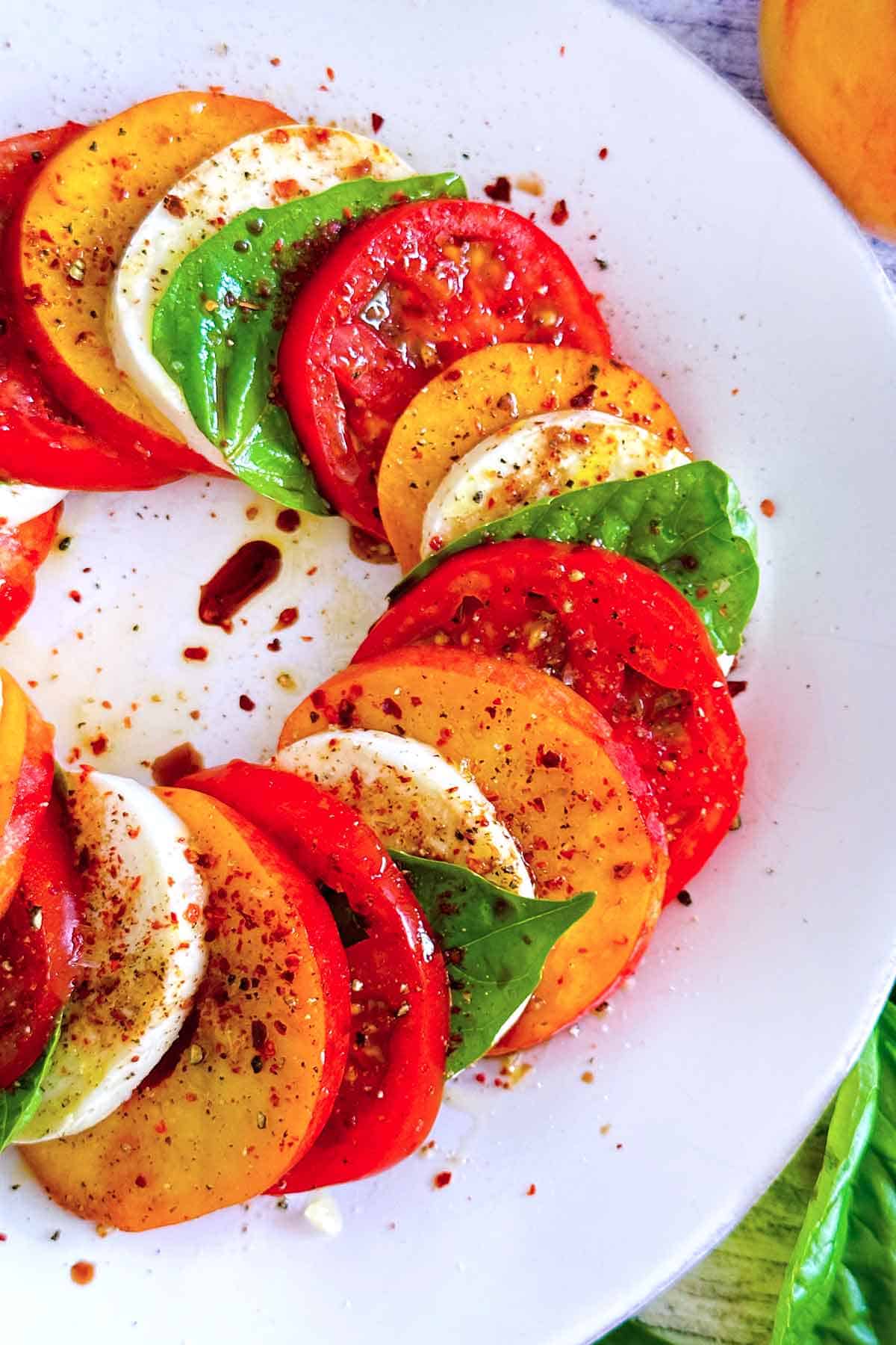 Close up photo of peaches, tomatoes, fresh mozzarella, and basil leaves layered on a plate for caprese salad, drizzled with balsamic vinegar and with spicy pepper on top.