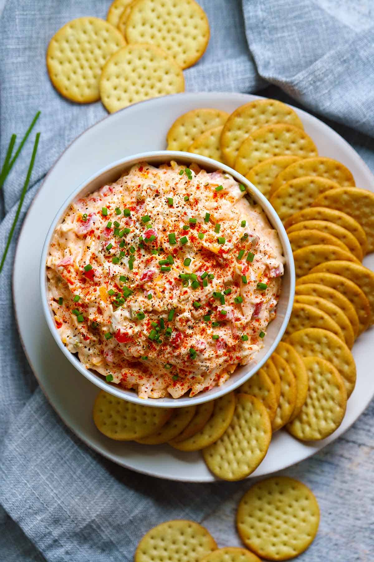 Overhead photo of pimento cheese in a white bowl garnished with chives and paprika, surrounded by crackers on a plate on a gray napkin background.