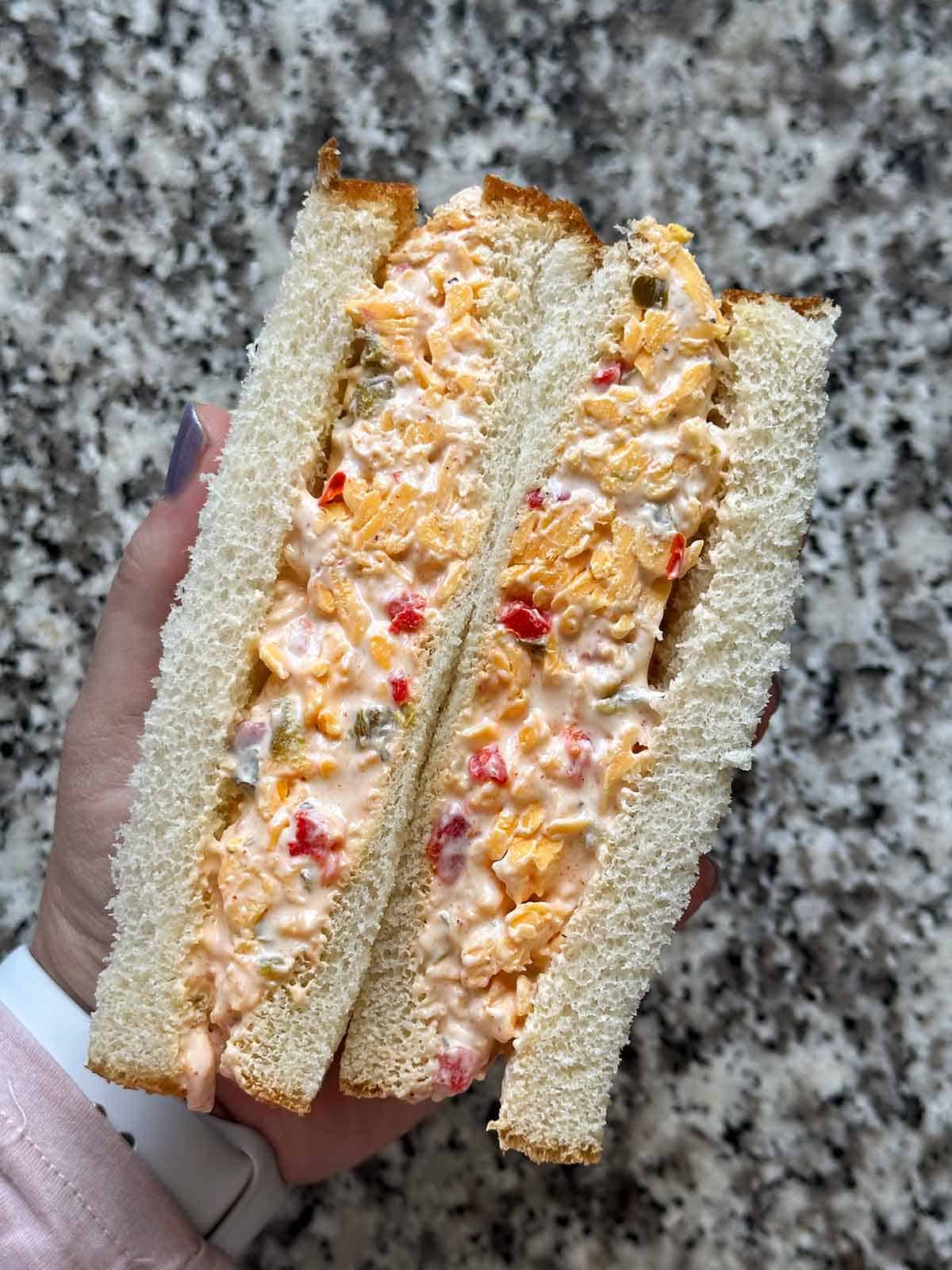 A hand holding a pimento cheese sandwich, cut in half, on white bread.