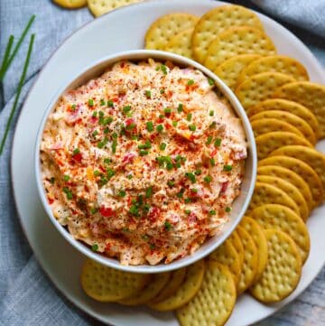 Square photo of overhead view of a bowl of pimento cheese surrounded by crackers on a plate.