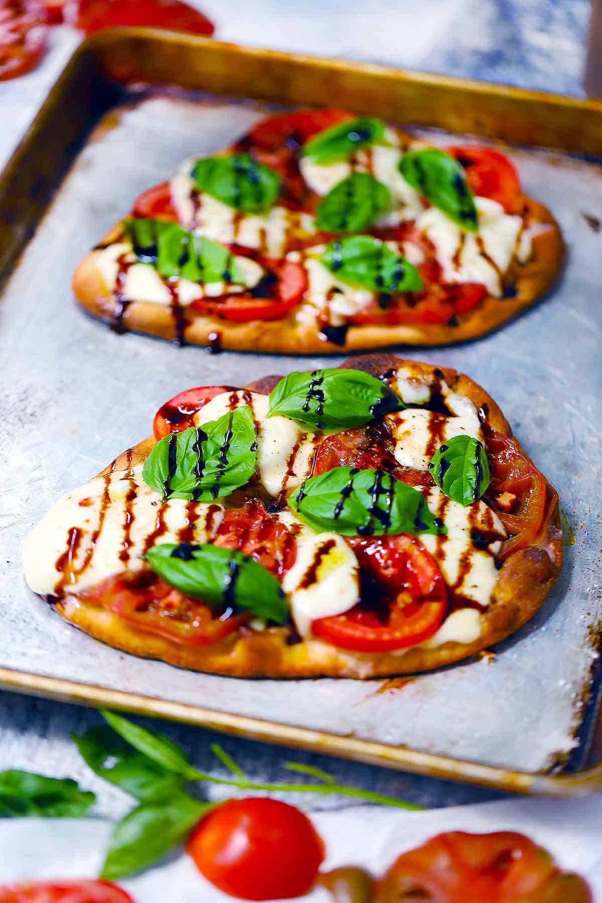 Two naan caprese pizzas on a baking sheet.