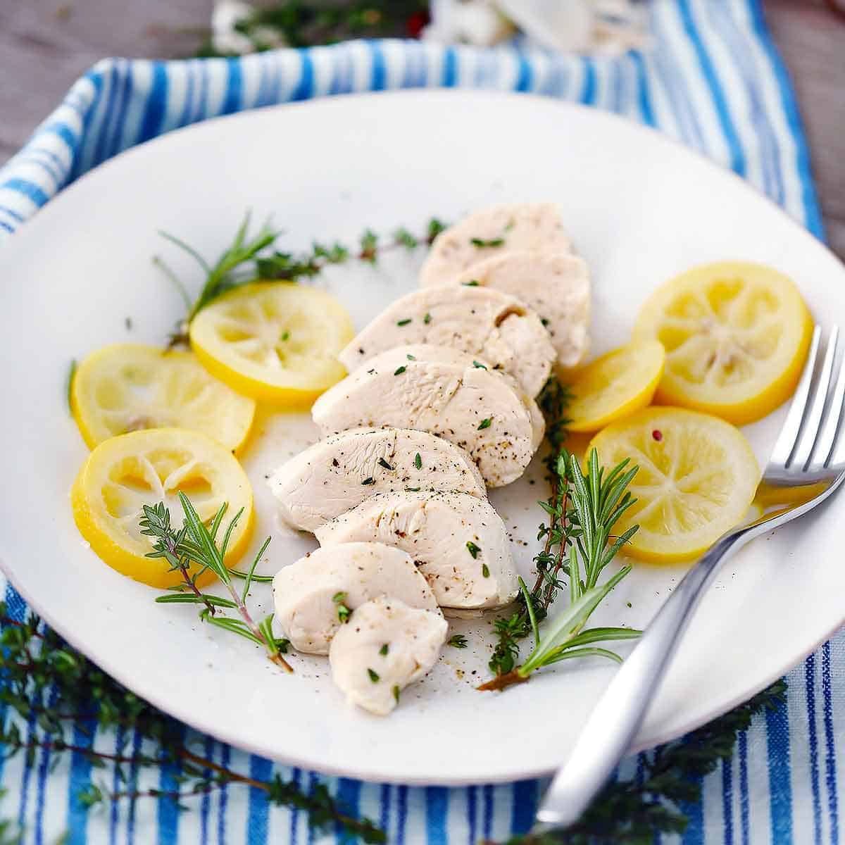 A white plate with a poached chicken breast sliced and herbs and lemon slices around it with a fork.