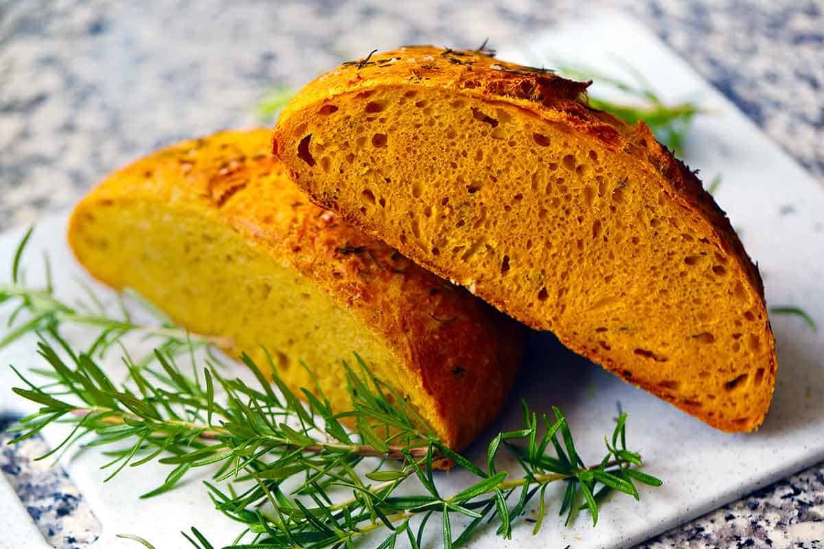 Crusty pumpkin no knead bread cut in half on a cutting board with sprigs of fresh rosemary.