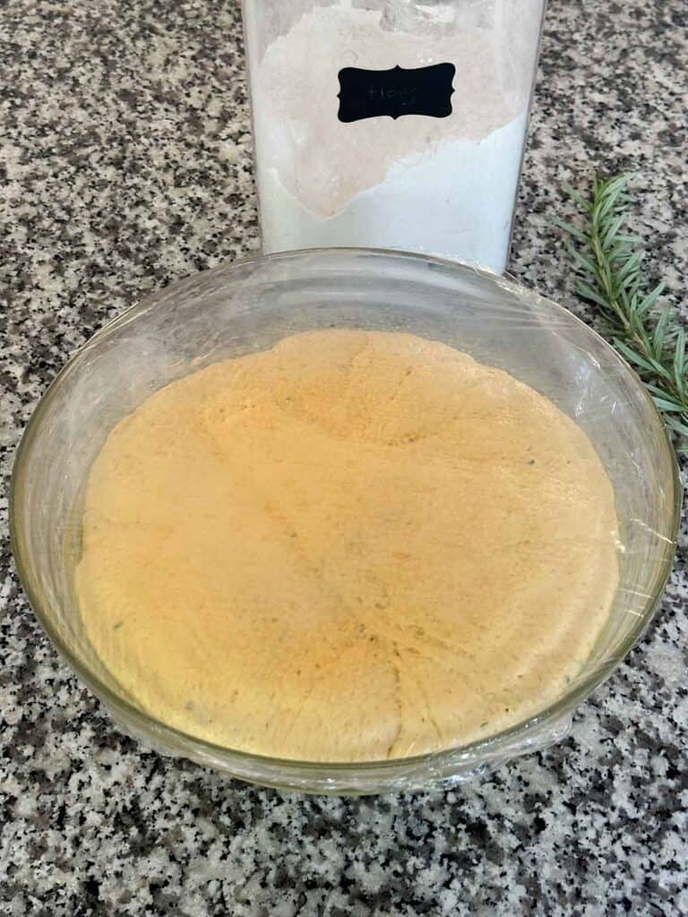A bowl with pumpkin bread dough in it after rising from the yeast, with plastic wrap covering it.