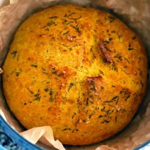 Square photo of a loaf of pumpkin no knead bread in a parchment lined Dutch oven, with rosemary and sea salt on top.